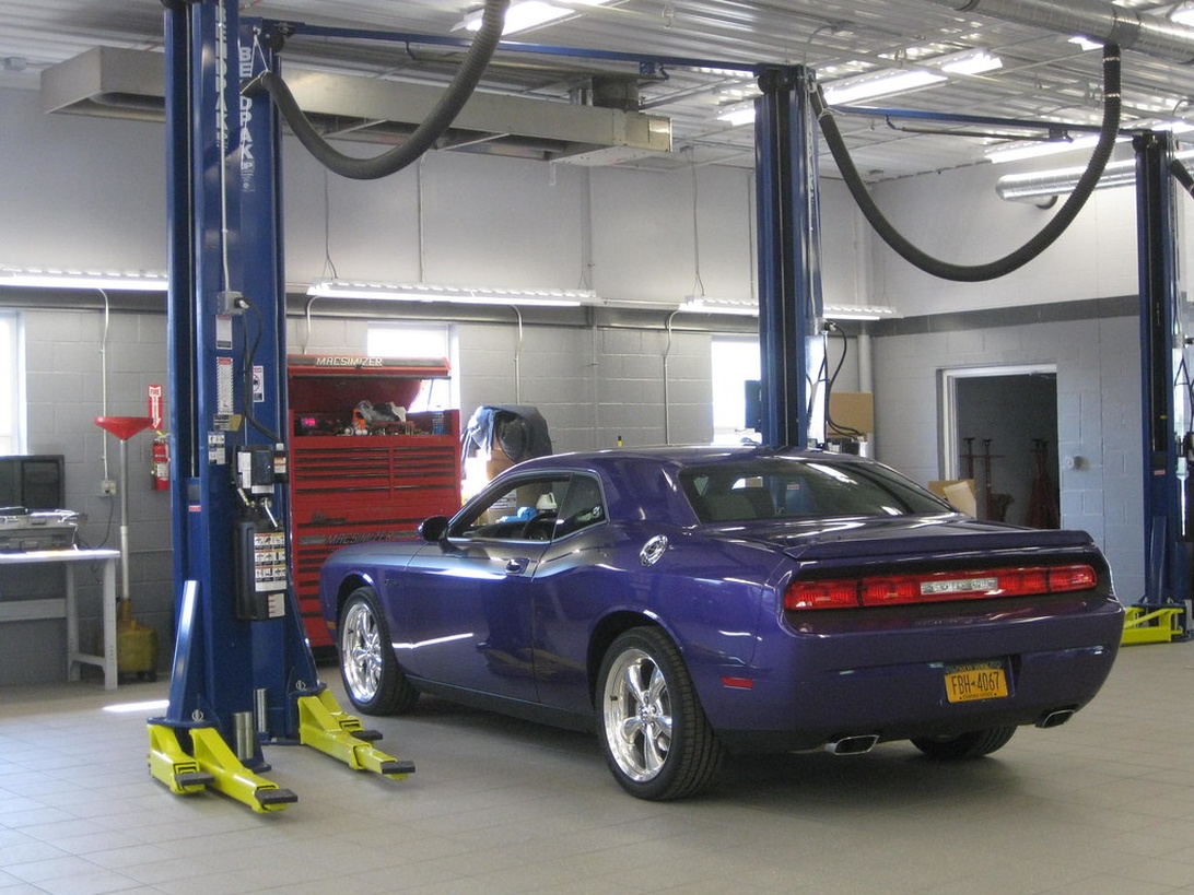 En modern bilserviceplats med blå hydrauliska lyftar och en lila Dodge Challenger upphöjd för underhåll. Verkstaden har rent klinkergolv, stark belysning och professionell utrustning inklusive verktygsskåp. Bilden visar tydligt en samtida bilverkstad i drift.